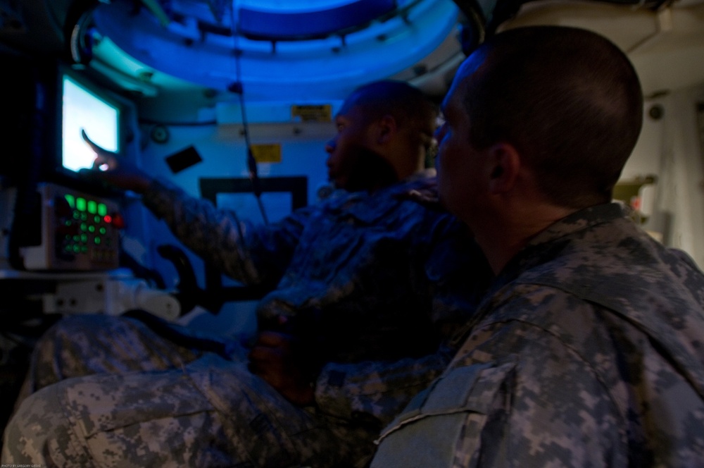 Two military personnel in U.S. Army ACU (Army Combat Uniform) camouflage are seated inside what appears to be a military vehicle or command post interior. One soldier is pointing at or operating a display panel with illuminated green and red indicator lights, while a monitor screen is visible in the background. The interior is lit with a distinctive blue ambient light, suggesting a low-light operational environment.