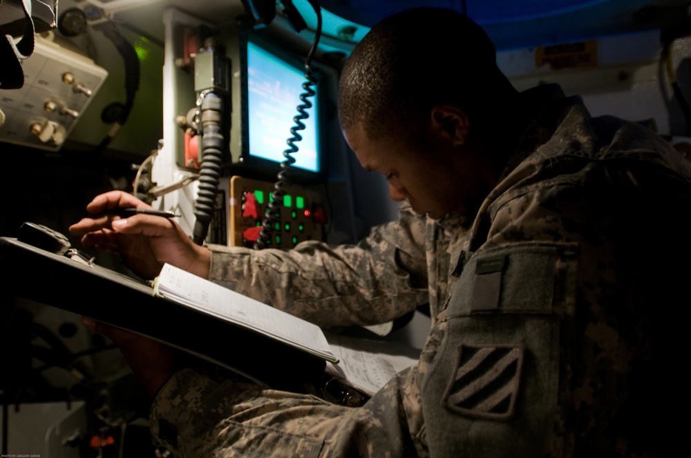 A U.S. Army soldier in ACU (Army Combat Uniform) is writing on a clipboard inside what appears to be a military command vehicle or operations center. The soldier wears a 3rd Infantry Division patch on the left shoulder. The environment is dimly lit with multiple electronic displays and communication equipment visible in the background.