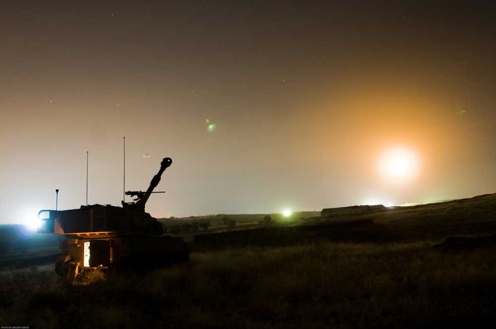 A nighttime military field scene showing a self-propelled artillery vehicle silhouetted against an illuminated sky. The image was taken during what appears to be a live-fire exercise or operational deployment, with a bright light source on the horizon likely from an illumination round or distant light source. A photographer credit reading 'Photo by Gregory Geske' is visible in the lower left corner.