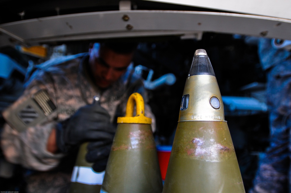 A military service member in camouflage uniform and black gloves works in the background, while two artillery projectiles are prominently displayed in the foreground. The projectiles appear to be olive-drab colored munitions, with one bearing a yellow safety cap and the other featuring a fuze assembly with visible markings. The photo credit visible in the lower left corner reads 'Photo by Gregory Gieske'.