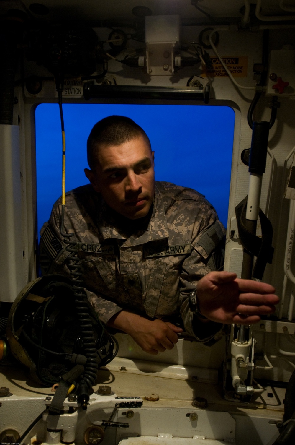 A U.S. Army soldier in ACU (Army Combat Uniform) is leaning through or into a hatch opening of what appears to be a military vehicle or simulator. The soldier, identified by the name tape 'CRUZ' on the uniform, appears to be inspecting or adjusting equipment inside the vehicle interior. The photo was taken by Gregory Gieske, as noted in the bottom-left watermark.