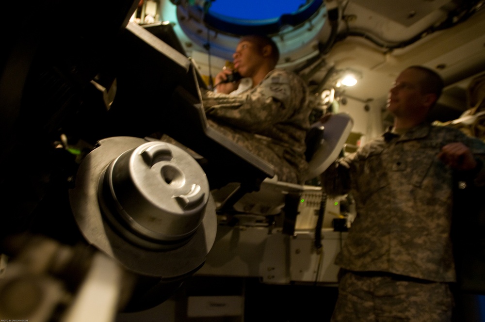 The image shows the interior of a military vehicle or command post with at least two soldiers in U.S. Army camouflage (ACU pattern) uniforms. One soldier appears to be speaking on a handset/radio while seated, and another stands nearby holding what appears to be a clipboard or document. A large mechanical component, possibly part of a weapon system or turret mechanism, is prominently visible in the foreground.