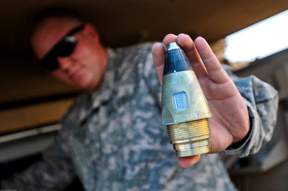 A U.S. Army soldier in ACU (Army Combat Uniform) holds up a artillery fuze component toward the camera for display or inspection. The object is held in sharp focus in the foreground while the soldier remains blurred in the background. The photo credit visible in the lower left reads 'Photo by Gregory Gieske'.
