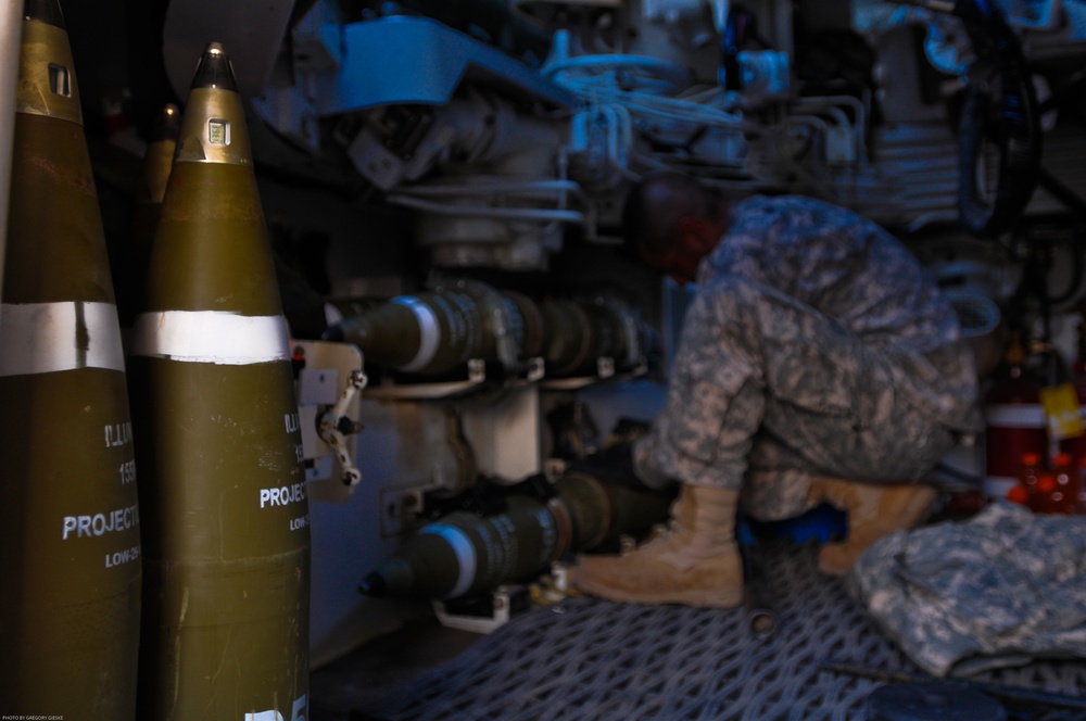 A soldier in US Army ACU (Army Combat Uniform) digital camouflage is working inside what appears to be a self-propelled artillery vehicle or armored vehicle interior. Two large artillery projectiles labeled 'PROJECTILE' and 'LOW-S' (likely Low-Smoke or Low-Smoke/Low-Drag) are prominently visible in the foreground. The image was taken by photographer Gregory Gieske.