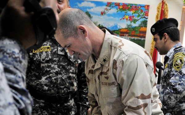 A U.S. Coast Guard officer wearing desert camouflage uniform with a one-star general insignia is leaning over a table examining plaques and awards at what appears to be a formal ceremony. The event involves personnel from multiple military/police organizations, including Iraqi security forces in digital camouflage uniforms. The table displays several commemorative plaques with gold circular medallions and an Arabic-language certificate.