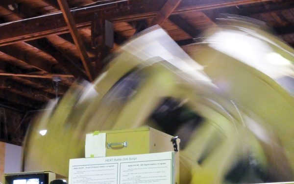 A military training environment showing a large rotating HEAT (Helicopter Emergency Egress Training) simulator device painted olive/yellow-green. A soldier in camouflage uniform operates a control panel on the left side of the frame. The large cylindrical/spherical apparatus is visibly spinning, as evidenced by the motion blur captured in the photograph.