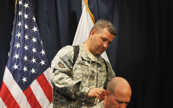 Two U.S. Army soldiers in ACU (Army Combat Uniform) camouflage are shown in what appears to be an official setting with an American flag and a white flag in the background. The standing soldier, identified by name tape as 'LUSTER', appears to be assisting the seated soldier, identified as 'CAMPSEY', by adjusting or fitting an earpiece or microphone near his ear. The setting suggests a formal military briefing, press conference, or official ceremony.