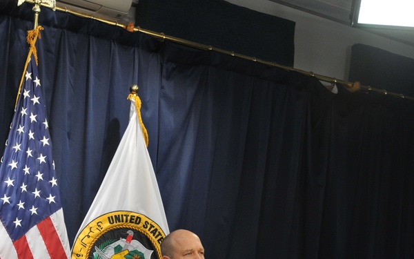 A U.S. Army soldier in ACU (Army Combat Uniform) is seated at a large formal wooden desk in what appears to be an official briefing or press conference setting. The name tape on the uniform reads 'CAMPSEY' and the uniform displays a 4th Infantry Division patch. Behind the soldier are a U.S. flag and a white organizational flag bearing a United States military command seal.