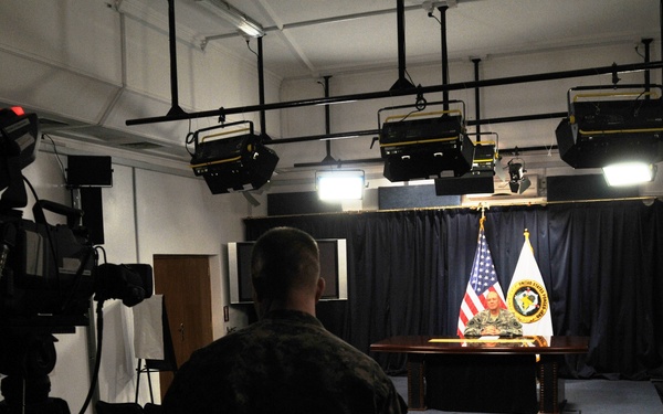 A military briefing or press conference is being filmed in a studio-like room with professional lighting rigs mounted on the ceiling. A uniformed military personnel sits at a desk on a small stage flanked by an American flag and what appears to be a military command seal/emblem flag. The scene is being recorded by at least one professional video camera operated by another uniformed individual in the foreground.