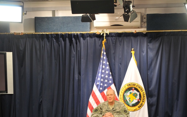 A U.S. Army officer in ACU (Army Combat Uniform) is seated at a formal wooden desk in what appears to be a military press briefing room or media studio. Behind him are two flags: the American flag and a United States Forces-Iraq (USF-I) organizational flag. The room is equipped with professional studio lighting and has rows of empty chairs facing the desk.