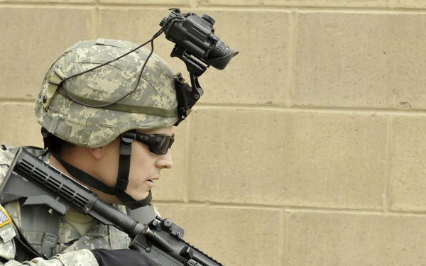 A U.S. Army soldier in full combat gear is positioned against a concrete block wall in what appears to be a training or operational environment. The soldier is holding an M4 carbine rifle with a rail system and a blank firing adapter attached to the muzzle, indicating a training exercise. The soldier is equipped with ACU (Army Combat Uniform) pattern camouflage, body armor, night vision device mount, and tactical accessories.