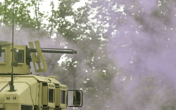 A tan/desert-colored military HMMWV (Humvee) is positioned on a gravel road surrounded by purple/violet colored smoke, likely from a smoke grenade or training device. The vehicle appears to be equipped with a turret-mounted weapon system on top. The setting appears to be a training exercise in a wooded or semi-rural environment.