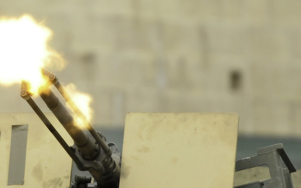 The image shows a multi-barrel automatic cannon mounted on a military vehicle, actively firing as evidenced by the muzzle flash and flame visible at the barrel tips. The weapon system appears to be mounted on a desert-tan colored armored platform with protective shield panels. The firing is captured mid-discharge with bright orange and yellow flames erupting from the muzzle.