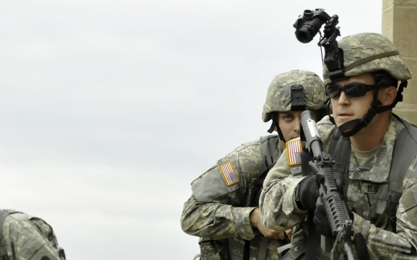 Three U.S. Army soldiers in full combat gear are conducting what appears to be a military training exercise near a stone or concrete block structure. The soldiers are wearing Army Combat Uniforms (ACU) in Universal Camouflage Pattern and are armed with M4/M16-style rifles. Two soldiers are taking a tactical position at the corner of a building while a third moves in the background.