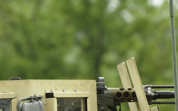 A U.S. Army soldier in ACU (Army Combat Uniform) is positioned in the turret of a tan/desert-colored military vehicle, likely a HMMWV (Humvee) or similar tactical vehicle. The vehicle is equipped with a mounted weapon system, appearing to be an M2 .50 caliber machine gun or similar crew-served weapon. The setting appears to be a training environment or exercise, with green foliage visible in the background.