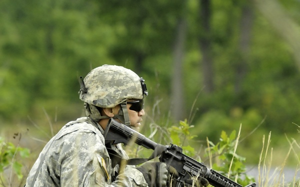A U.S. Army soldier in Army Combat Uniform (ACU) digital camouflage is positioned in tall grass and vegetation, holding an M4 carbine rifle in a ready stance. The soldier is wearing a combat helmet, ballistic sunglasses, and tactical gloves while appearing to scan or cover a sector. The setting appears to be a field training exercise based on the yellow training muzzle cap visible on the rifle barrel.
