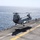 A U.S. Marine Corps CH-46 Sea Knight tandem-rotor helicopter is hovering just above the flight deck of a naval amphibious assault ship at sea. The helicopter appears to be in the process of landing or taking off, with its shadow clearly visible on the gray deck below. A flight deck crew member in a yellow vest is visible on the right side of the deck.