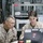Two U.S. Marines in desert MARPAT camouflage uniforms are seated together reviewing content on an HP laptop computer. The individual on the left has a name tape reading 'MCMILLIAN' and wears glasses along with a gold device on their chest, suggesting senior rank. They appear to be in a military operations or communications room with networking equipment, cables, and printers visible in the background.