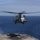 A large military helicopter is hovering just above the flight deck of a naval vessel over open ocean. The helicopter appears to be in the process of landing or departing from the ship's deck. The image was taken from the ship's deck level, providing a direct frontal view of the aircraft.