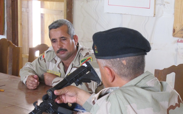 Two military personnel in desert camouflage uniforms are seated at a wooden table in what appears to be an indoor briefing or training room. One soldier wearing a black beret is handling a disassembled rifle, while the other soldier with rank insignia and unit patches observes. The setting suggests a weapons familiarization or maintenance training session.