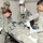 Military medical personnel in U.S. Army ACU (Army Combat Uniform) are treating a patient on a field stretcher inside what appears to be a military medical tent or aid station. At least four soldiers are visible, with one applying bandaging while wearing blue medical gloves, and another holding a clipboard. The scene depicts a combat casualty care or medical training exercise.