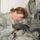 A female U.S. Army soldier in ACU (Army Combat Uniform) is leaning over and attending to what appears to be a patient or simulated casualty in what looks like a medical treatment tent or field hospital. She appears to be performing a medical assessment or treatment procedure. A second soldier in full combat gear, including a radio and stethoscope, stands nearby.