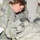 A female U.S. Army soldier in Army Combat Uniform (ACU) pattern is leaning over and attending to another soldier who appears to be lying on a litter or stretcher. The standing soldier appears to be performing a medical assessment or providing care, with a clipboard visible to her left. The setting appears to be inside a military tent or field medical facility.