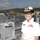 A female U.S. Navy officer in white service dress uniform stands on the deck of a naval vessel, smiling at the camera. The ship appears to be docked at a harbor with a waterfront town visible in the background. The vessel is decorated with colorful signal flags strung across the ship, suggesting a ceremonial or public event.