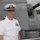 A U.S. Navy officer in white service dress uniform poses on the deck of a naval warship, standing in front of a large deck gun turret. The setting appears to be a port visit or public event, as civilians and other personnel are visible in the background. The waterfront with trees and buildings is visible in the distance.