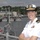 A female U.S. Navy officer in white service dress uniform stands on the deck of a naval vessel at a harbor installation. She is smiling with arms crossed, wearing a combination cover with gold officer insignia and displaying ribbons and a gold device on her uniform. Multiple naval ships and signal flags are visible in the background at what appears to be a naval base.