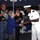 A formal ceremony appears to be taking place outdoors at dusk under a tent, likely on or near a naval vessel. A senior Naval officer in white dress uniform is shaking hands with an elderly woman in a blue jacket, appearing to present her with an award or recognition. Several onlookers, including civilians and other naval personnel, are applauding in the background.