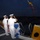 Two naval officers in white dress uniforms are standing on the deck of a ship at dusk or dawn, rendering a salute. The vessel is decorated with international maritime signal flags along its rigging and railing. A small group of additional personnel is visible in the background on the left side of the deck.