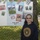 A woman wearing a United States Navy hoodie stands in front of a poster board attached to a chain-link fence. The poster is a collage celebrating Fleet Week 2010, featuring labeled sections for Army, Air Force, Navy, Marines, and Coast Guard with various photographs. The setting appears to be an outdoor event, likely a school or community gathering.