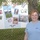 A woman with red hair wearing a light blue 'NAVY' t-shirt stands smiling in front of a poster board attached to a chain-link fence. The poster is a Fleet Week 2010 display featuring photos and labels for five military branches: Army, Air Force, Navy, Marines, and Coast Guard. The setting appears to be an outdoor event, likely a community or school gathering.