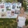 A woman stands smiling in front of a poster board attached to a chain-link fence at Fleet Week 2010. The poster displays photos representing all five U.S. military branches — Army, Air Force, Navy, Marines, and Coast Guard — along with family photos and a sponsor logo. The woman is wearing a white tank top and a red, white, and blue ribbon pin.