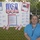 A woman wearing a blue polo shirt and lanyard stands in front of a patriotic USA-themed poster board attached to a chain-link fence. The display features a metallic 'USA' balloon letters decoration, a central text titled 'A Mother's Prayer,' and numerous small note cards arranged around the board. The setting appears to be an outdoor event or memorial display, likely military or patriotic in nature.