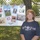 A middle-aged woman with reddish hair stands in front of a chain-link fence, posing next to a poster board display. The poster is a collage celebrating 'Fleet Week 2010' featuring labels for all five U.S. military branches: Army, Air Force, Navy, Marines, and Coast Guard. The setting appears to be an outdoor community or school event on a sunny day.