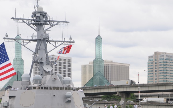 A U.S. Navy Arleigh Burke-class guided-missile destroyer bearing hull number 102 is docked or maneuvering in an urban waterway, assisted by a FOSS tugboat named 'Pacific Escort'. Sailors in white dress uniforms are manning the rails along the ship's deck, a traditional naval ceremony. The city skyline in the background, featuring distinctive green glass spires, is consistent with Portland, Oregon.