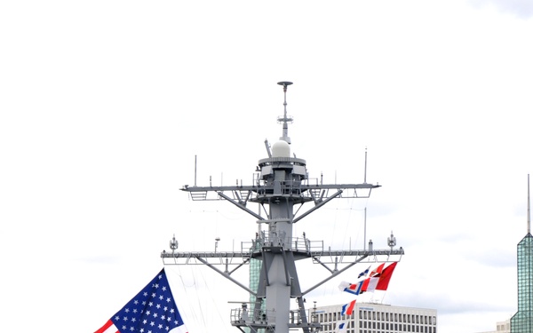 A U.S. Navy Arleigh Burke-class guided-missile destroyer bearing hull number 102 is shown entering or docking at a port, likely Portland, Oregon based on the visible urban skyline and bridge. The ship is dressed with a large American flag and signal flags, with sailors in white dress uniforms manning the rails in a formal 'manning the rails' ceremony. Several civilian and escort vessels are visible in the surrounding waterway.
