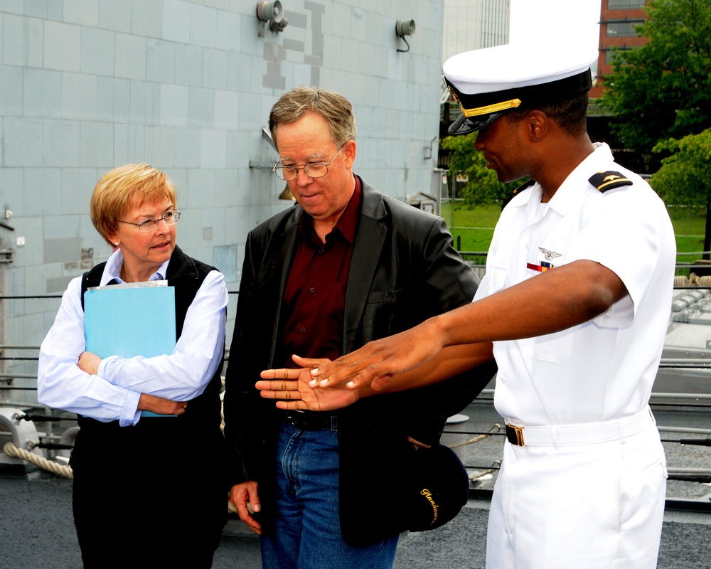 A U.S. Navy officer in white dress uniform is showing or explaining something in his open palm to two civilian visitors, a man and a woman, on what appears to be the deck of a naval vessel. The officer is gesturing with an outstretched hand while the civilians look attentively at what he is presenting. The setting appears to be an official tour or visit aboard a warship.