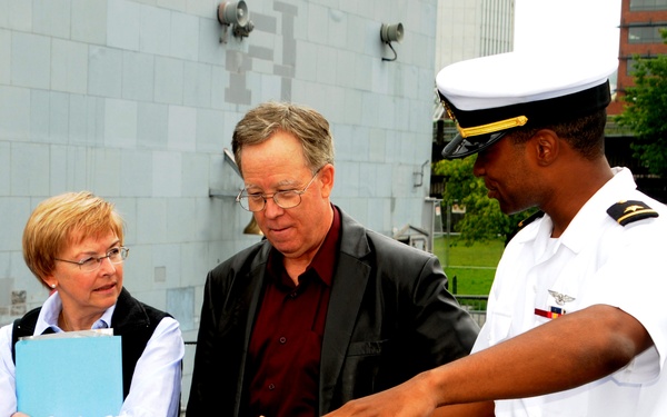 A U.S. Navy officer in white dress uniform is showing or explaining something in his open palm to two civilian visitors, a man and a woman, on what appears to be the deck of a naval vessel. The officer is gesturing with an outstretched hand while the civilians look attentively at what he is presenting. The setting appears to be an official tour or visit aboard a warship.