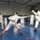 Two fencers in full white protective gear and masks are engaged in an active bout on a blue gymnasium floor. A referee or coach in casual white clothing observes from the left side, while additional individuals are visible in the background. The setting appears to be an indoor sports hall equipped with fencing equipment hanging on the right wall.
