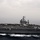 A large US Navy aircraft carrier, hull number 75 visible on the island superstructure, is underway at sea under overcast skies. The flight deck is populated with numerous fixed-wing aircraft and helicopters in various positions. The vessel is producing a visible white wake, indicating active forward movement through calm open ocean.