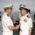 Two senior naval officers in white dress uniforms are shaking hands on the deck of a naval vessel at sea. The officer on the right displays a large array of service ribbons and rank insignia consistent with a senior U.S. Navy captain, while the officer on the left wears a heavily gold-braided cap suggesting flag-rank or senior officer status, possibly from a foreign navy. A junior enlisted sailor in traditional white uniform stands in the background.