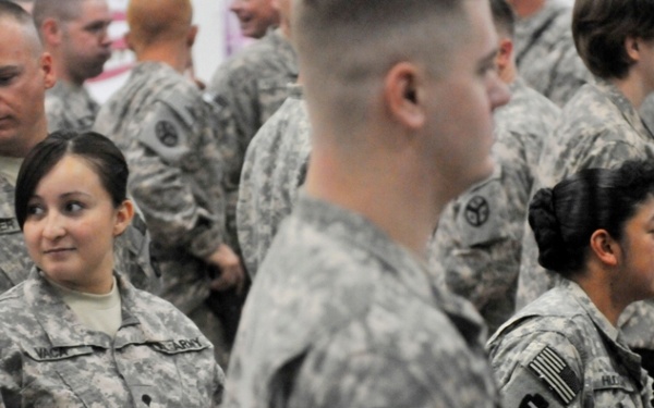 A group of U.S. Army soldiers in ACU (Army Combat Uniform) digital camouflage pattern are gathered in what appears to be an indoor assembly or formation area. Multiple soldiers are visible, including at least two female soldiers, with unit patches and American flag patches visible on their sleeves. The scene suggests a pre-deployment ceremony, unit formation, or military gathering event.