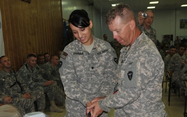 Two U.S. Army soldiers in ACU (Army Combat Uniform) are cutting a large ceremonial cake decorated in blue and white with a star and swirl design. The event appears to be a military celebration or unit anniversary, taking place in a dining facility (DFAC) with numerous other soldiers seated and watching. A U.S. Army flag dated '1775' is visible in the background.
