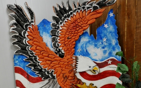 The image shows a U.S. Army birthday celebration setup featuring a decorated cake commemorating the Army's 235th birthday on June 14, 2010. A large painted wooden cutout of a bald eagle with an American flag backdrop serves as a patriotic display behind a yellow satin-draped table. The foreground table, covered in blue cloth, holds the celebration cake along with serving containers and a cake knife.