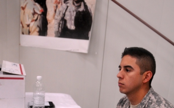 A US Army soldier in ACU (Army Combat Uniform) is operating audio/DJ equipment at a table covered with a white cloth. The soldier appears to be working with a Native Instruments controller and a mixing board, suggesting a music production or DJ setup. A shoulder patch with a 'T' insignia is visible on the uniform.