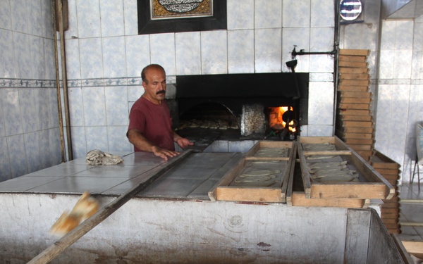 A traditional flatbread bakery showing two men working in a tiled room with a wood-fired oven. One man is actively working at a large work surface sliding bread toward the oven, while another man stands observing. Baked and unbaked flatbreads are visible at various stages of production.