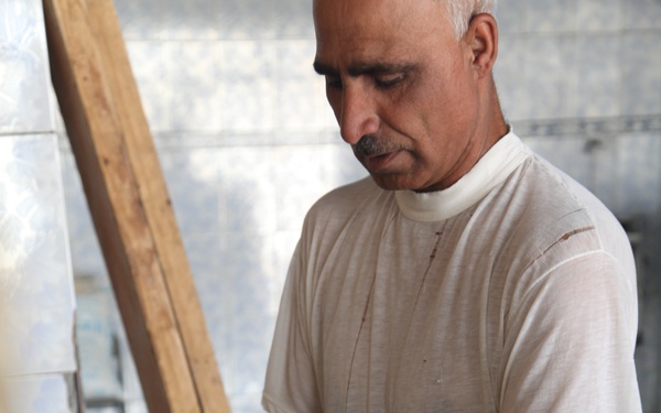 A middle-aged man is working in what appears to be a traditional bakery, handling flatbread dough on a wooden rack or tray. He is wearing a white t-shirt and is focused on stretching or placing dough onto a slatted wooden surface. The setting appears to be a small, informal bread-making workshop.